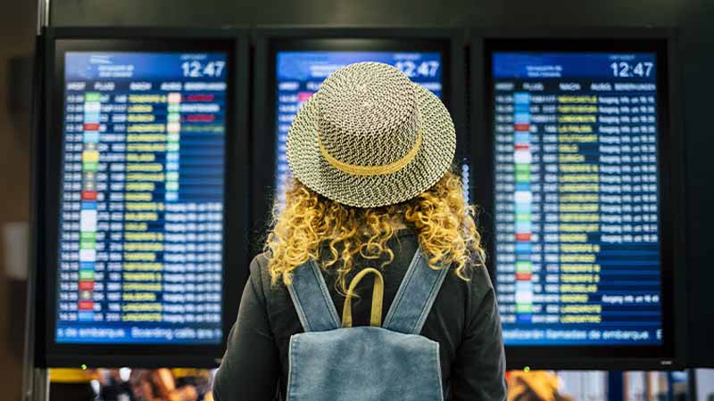 Mulher com chapéu e mochila de costas consulta os painéis dos voos ao fundo