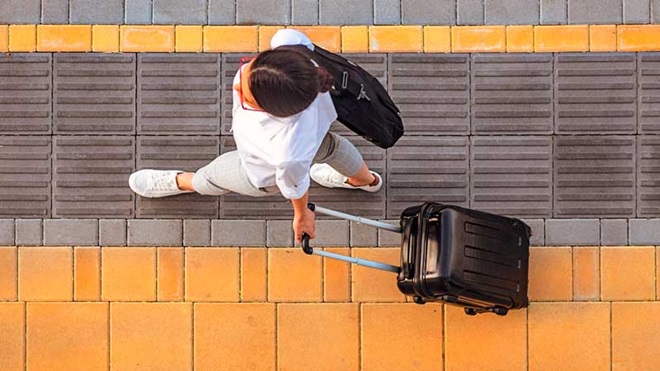Vista de cima de uma mulher vestida de branco a andar puxando um trolley de mala de viagem preta - alusão a ida de férias