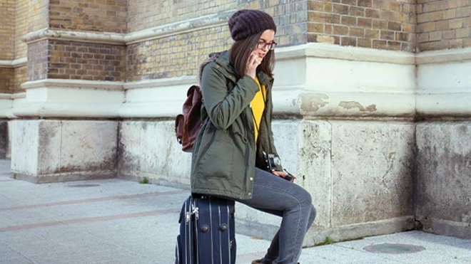 Mulher jovem ao telemóvel, sentada em cima de uma mala de viagem, na rua