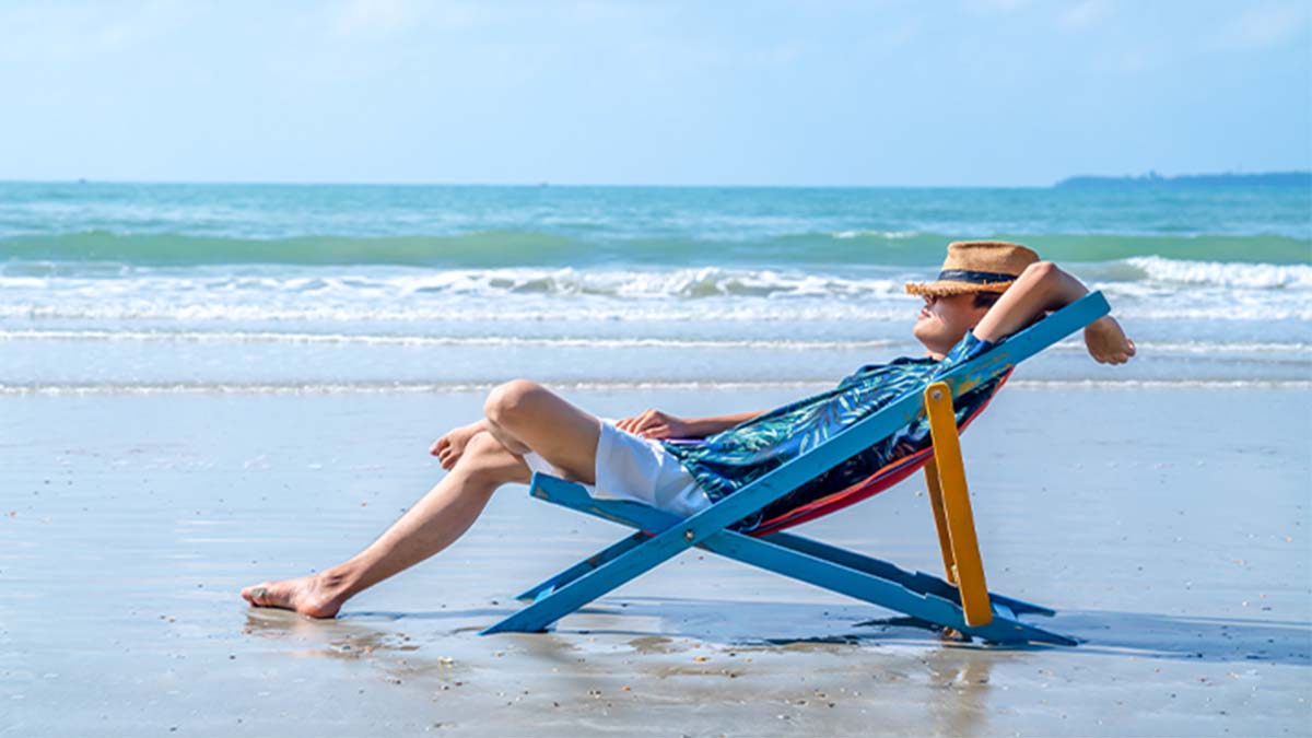 Pessoa reclinada numa espreguiçadeira de madeira e tecido azul, colocada na areia junto ao mar, com chapéu de palha, ondas suaves e céu azul ao fundo.