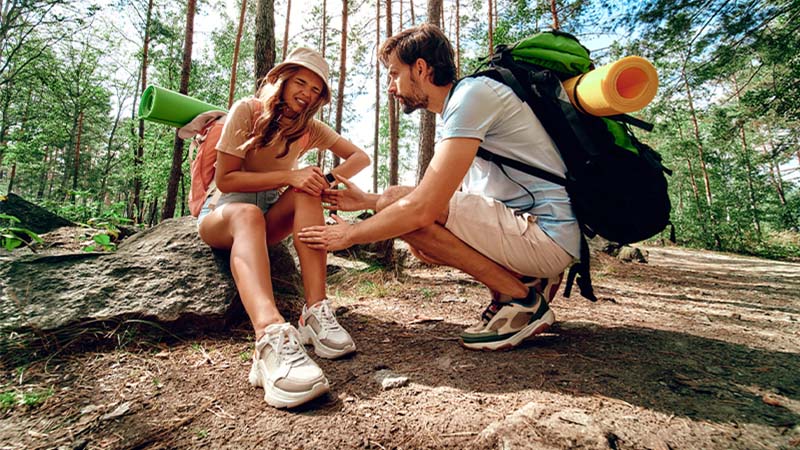 Casal em férias na natureza, com mochilas, mulher lesionada no joelho e homem a dar apoio