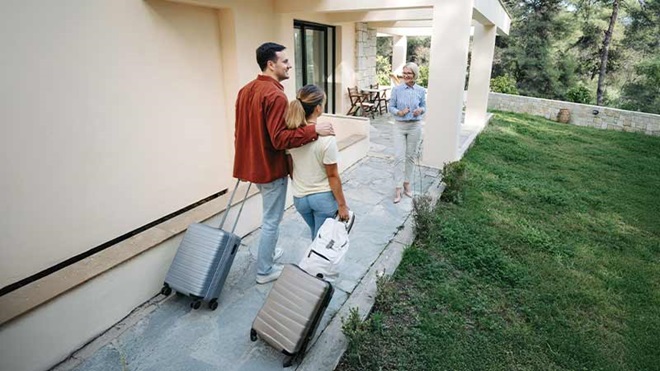 Casal a chegar a uma casa de férias com relvado, com malas de viagem e a senhoria a recebê-los