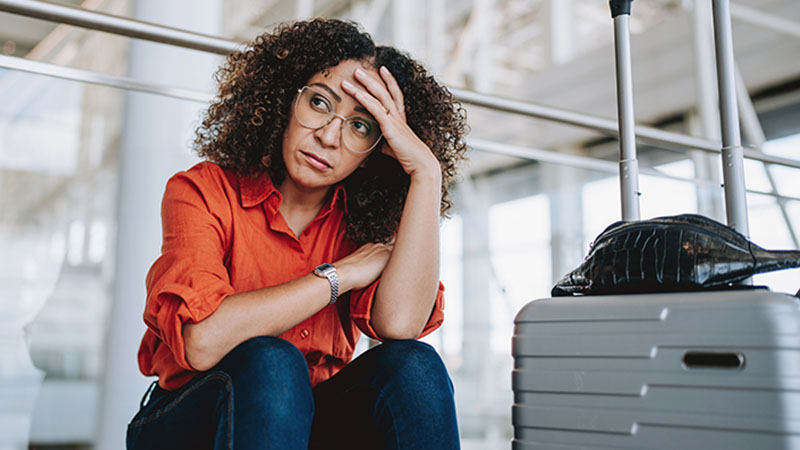 Pessoa sentada num aeroporto, com a cabeça apoiada na mão, ao lado de uma mala rígida cinzenta; cadeiras metálicas e estrutura do terminal ao fundo.