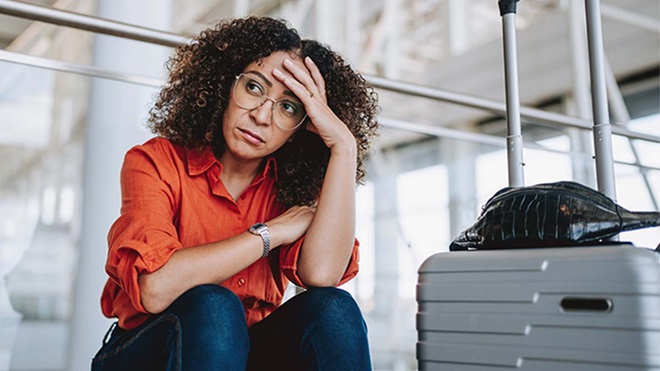 Pessoa sentada num aeroporto, com a cabeça apoiada na mão, ao lado de uma mala rígida cinzenta; cadeiras metálicas e estrutura do terminal ao fundo.