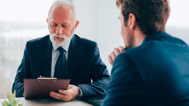Senhor idoso vestido de preto preenche documentos numa pasta, auxiliado por um jovem de fato escuro de costas