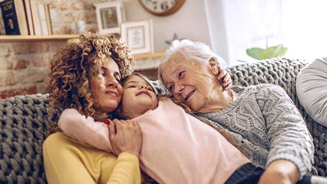 Três gerações: avó, com bastante idade, filha e neta, abraçadas no sofá da sala