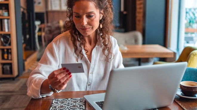 Mulher olha para o cartão bancário, sentada numa sala em frente a um portátil