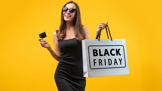 Mulher com vestido preto, cartão multibanco na mão, óculos de sol e um saco branco de papel a dizer black friday. Fundo amarelo