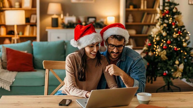 Casal na sala a fazer compras em laptop, com gorros de pai natal e árvore de natal 