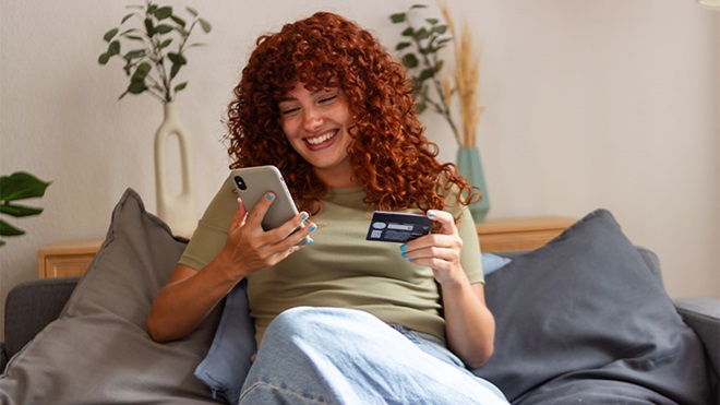 Mulher jovem a fazer compras online no telemóvel, com um cartão de crédito na mão