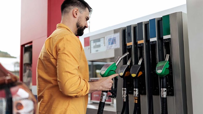 Pessoa abastece um veículo numa bomba de combustível, segurando uma mangueira verde, com várias bombas alinhadas e carros visíveis ao fundo.