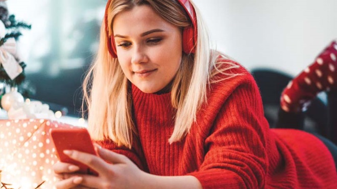 Jovem vestida com camisola de lã vermelha, em casa, junto à árvore e aos presentes, com headphones vermelhos e smartphone de capa vermelha na mão