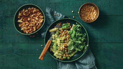 vista de mesa de madeira verde escura com 3 taças de diferentes tamanhos, uma grande com massa e verduras, outra média com nozes e frutos secos e a terceira pequena com (leguminosas).