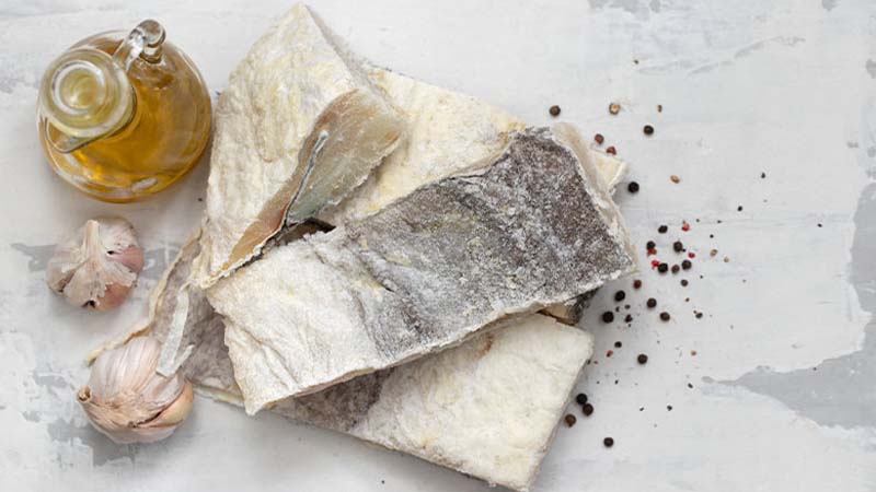 Postas de bacalhau salgado numa mesa branca, com uma cabeça de alhos e uma garrafa de azeite ao lado