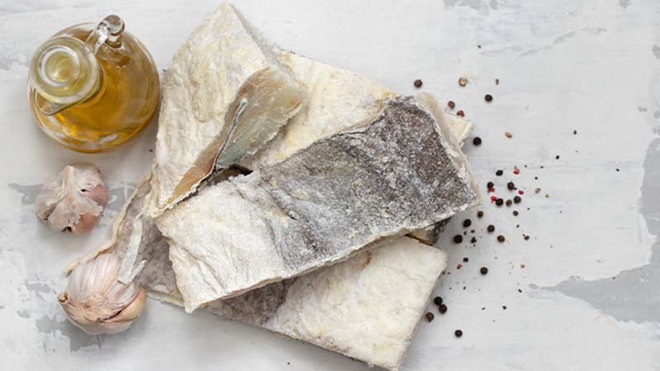 Postas de bacalhau salgado numa mesa branca, com uma cabeça de alhos e uma garrafa de azeite ao lado