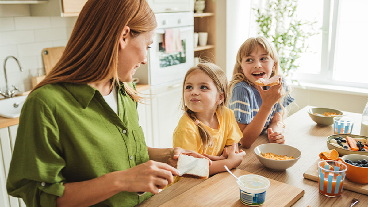 Pessoa adulta sentada à mesa de cozinha com duas crianças; tigelas de cereal; luz natural entra por uma janela ao fundo.