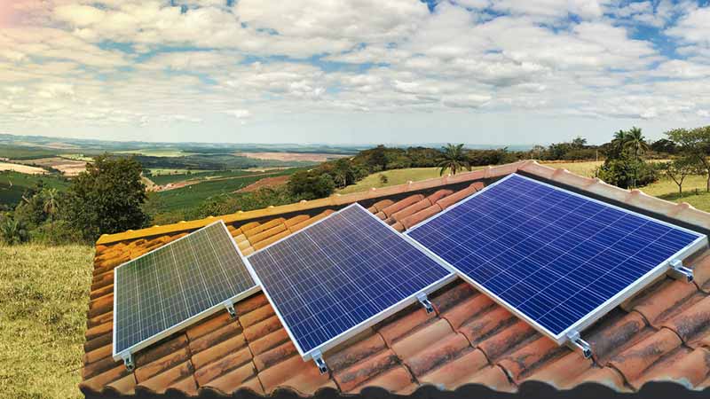 Pormenor de um telhado cor de tijolo com três painéis solares instalados, dispostos em escada, com uma paisagem verde como fundo.
