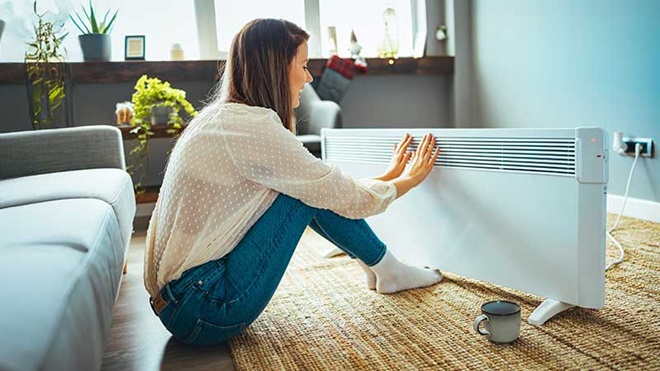 Pessoa com os pés estendidos em cima de um radiador de parede. Os pés têm meias calçadas. Ao lado há um móvel, em cima do qual está uma caneca de chá.