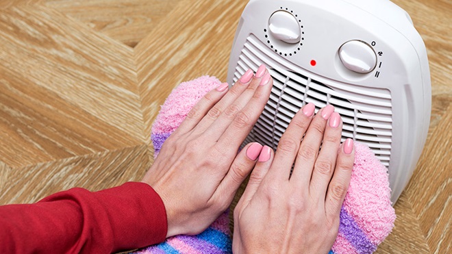 Plano aproximado de duas mãos pousadas sobre um par de pés protegidos com meias quentes, junto de um termoventilador em funcionamento, pousado sobre um chão de madeira.