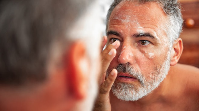 Homem sénior hidrata a pele em frente ao espelho - alusão a pele com dermatite atópica