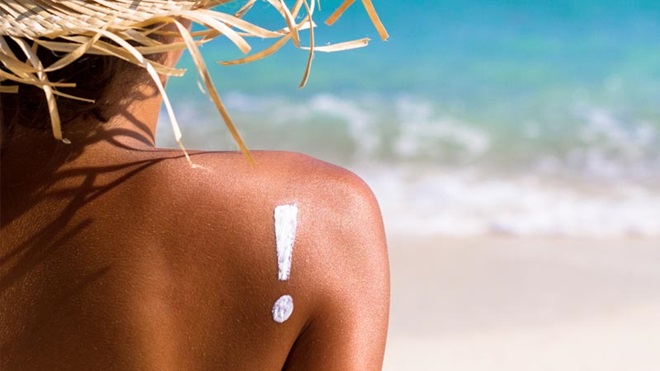 Mulher de costas com pele bronzeada em frente à praia; na zona do ombro, com creme branco tem um ponot de esclamação desenhado