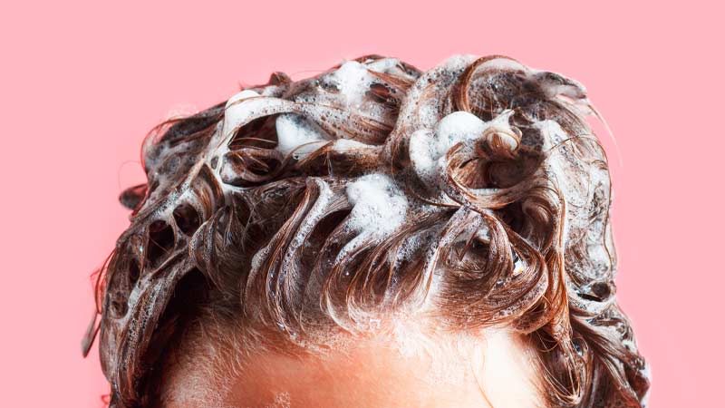 Grande plano do topo de uma cabeça de mulher a lavar o cabelo com champô, fundo cor-de-rosa