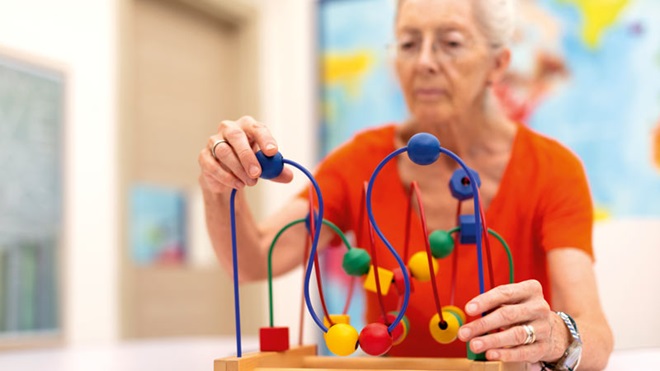 Idosa sentada em frente a uma mesa de madeira, tenta fazer um jogo de peças de madeira coloridas