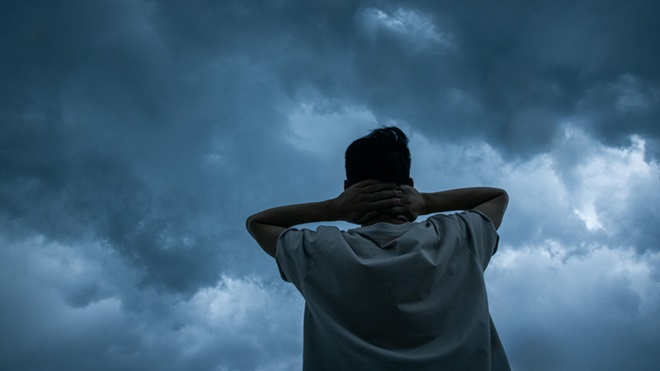 Homem de costas com as mãos no pescoço, céu de trovoada, carregado; ambiente que sugere preocupação