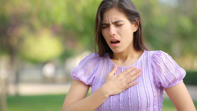 Pessoa ao ar livre, com uma mão sobre o peito, a usar um vestido lilás às riscas de manga curta, num ambiente exterior com vegetação desfocada ao fundo.