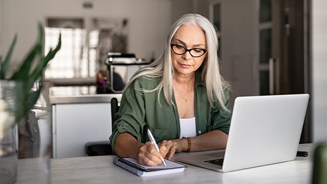 Mulher de meia idade a consultar informação num computador portátil, enquanto tira notas num bloco