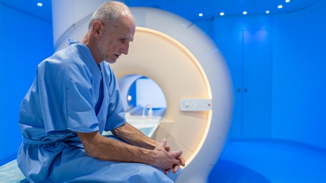 Homem sénior, vestido com bata de hospital aguarda sentado com ar preocupado numa sala azulada, junto de um equipamento médico de grandes dimensões (scanner médico)