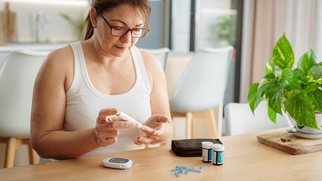 Mulher sentada à mesa, em casa, faz um teste de glicémia picando o dedo