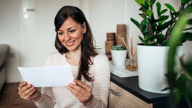 Pessoa sentada em casa a ler uma folha de papel, com plantas e objetos decorativos ao redor.