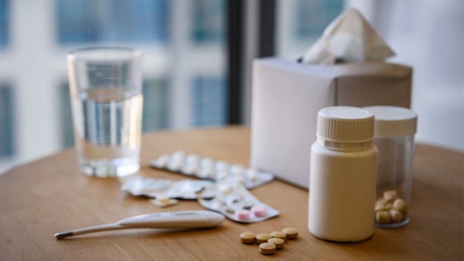 Vários medicamentos, lenços de papel, copo de água e termómetro em cima de mesa