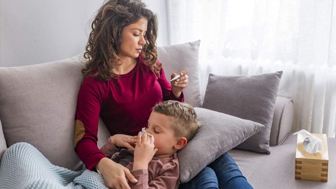 Mãe sentada no sofá com a cabeça do filho de 7 anos no colo; a crianºa assoa-se; a mãe vê a temperatura no termómetro. Ao lado está uma caixa de lenços de papel