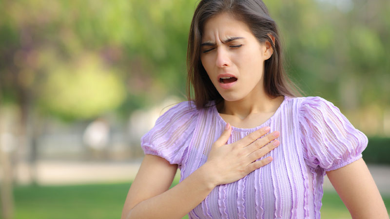 Pessoa ao ar livre, com uma mão sobre o peito, a usar um vestido lilás às riscas de manga curta, num ambiente exterior com vegetação desfocada ao fundo.