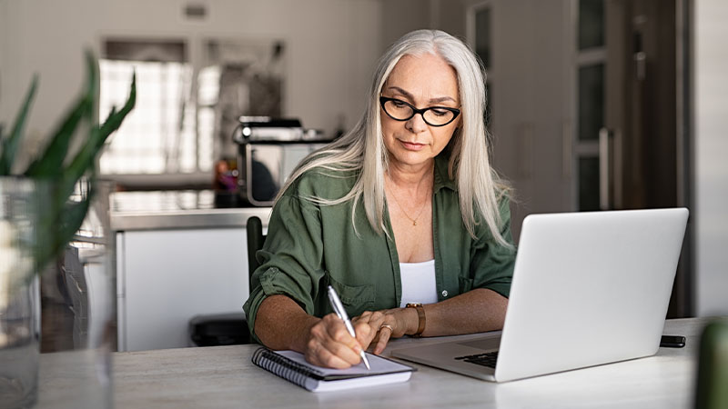 Mulher de meia idade a consultar informação num computador portátil, enquanto tira notas num bloco