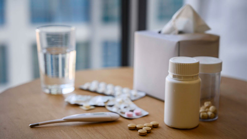 Vários medicamentos, lenços de papel, copo de água e termómetro em cima de mesa