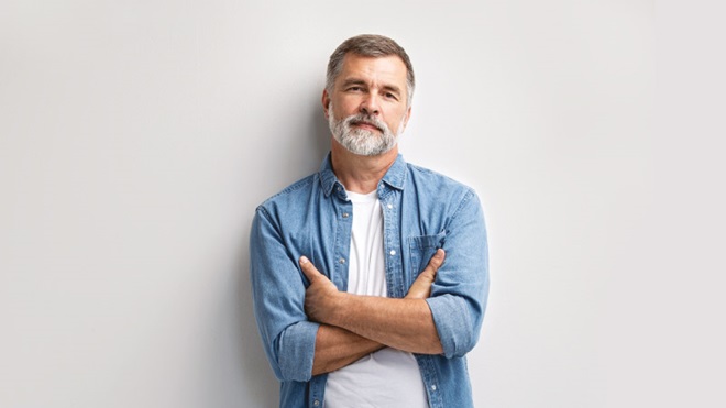 Plano médio de homem de meia idade, atraente, com barba, vestido com tshirt branca e camisa de ganga por cima, encostado a uma parede cinzenta. 