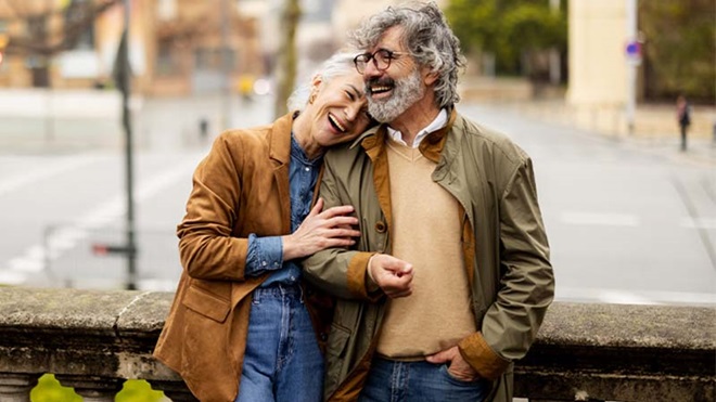 Casal de meia idade encostado a uma balaustrada num espaço urbano, ambos vestidos com casacos, enquanto caminham ou fazem uma pausa ao ar livre.