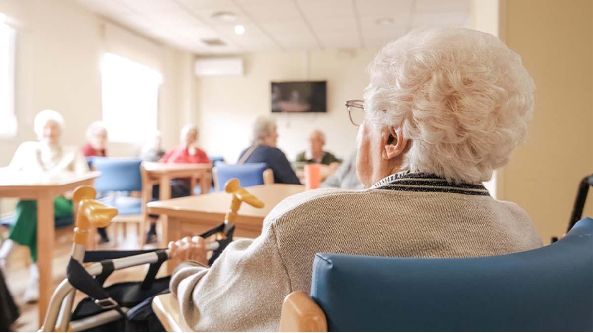 Idosos sentados numa sala comum de lar ou centro de dia, com mesas e cadeiras, enquanto uma pessoa em primeiro plano observa o ambiente a partir de uma cadeira de rodas.