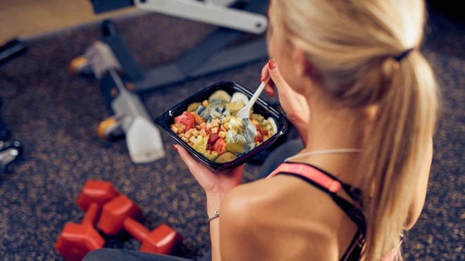 Mulher de costas com roupa desportiva a comer uma salada, com pesos e aparelho de fitness em pano de fundo
