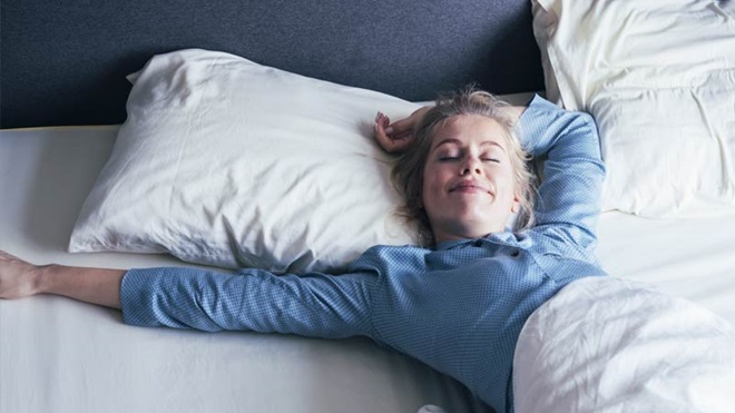 Mulher deitada numa cama com lençois brancos e pijama azul com um ar muito descansado, espreguiça-se