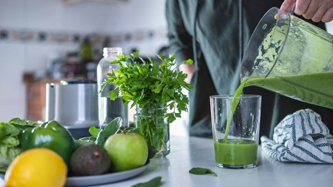 Pessoa a servir um smoothie num copo, ao lado de uma bandeja com frutos, legumes e molho de salsa