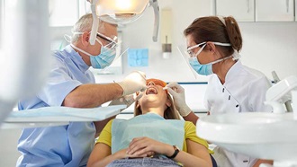 Mulher deitada na cadeira do dentista de boca aberta. Dentista à esquerda e assistente dentária à direita, realizam procedimento na mulher.