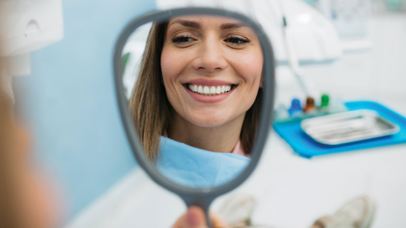 Grande plano de uma mulher jovem e atraente a sorrir ao espelho com uns dentes perfeitos; à volta um cenário desfocado de um consultório dentário