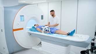 Paciente num aparelho de grandes dimensões, faz um procedimento médico num hospital, com um enfermeiro ao lado