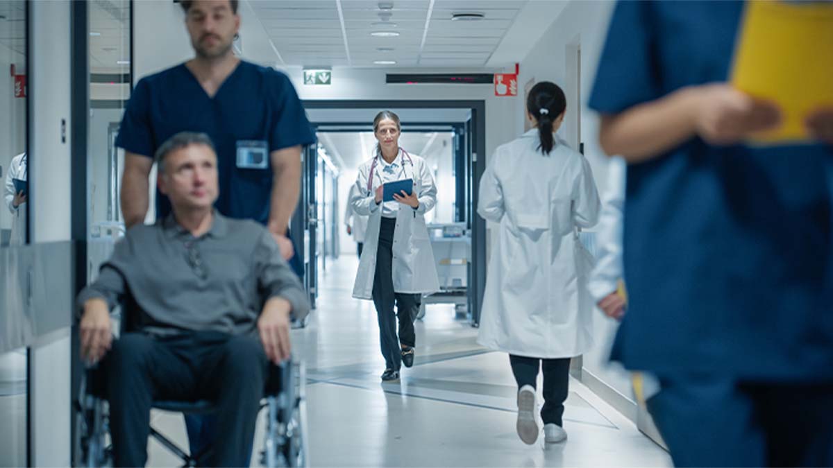 Corredor de hospital com pessoa numa cadeira de rodas empurrada por profissional de saúde, enquanto outros profissionais circulam ao fundo entre portas e equipamentos clínicos.