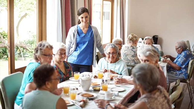 idosos sentados numa mesa num lar tomam o pequeno-almoço acompanhados por uma cuidadora