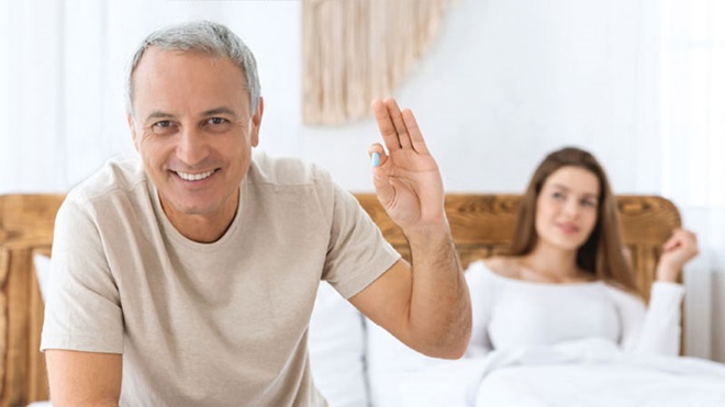 Homem novo em primeiro plano, sentado numa cama segura um comprimido azul; no plano de fundo, na cama, um pouco desfocada, está uma mulher